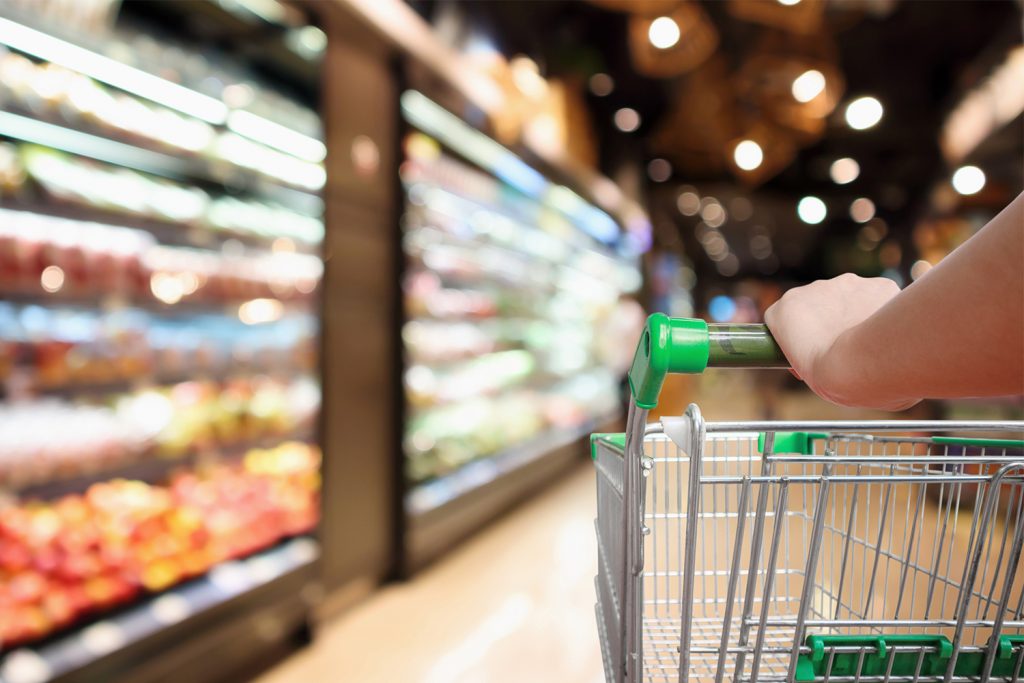 A person pushing a shopping cart down an aisle in a brightly lit grocery store. Shelves on the left side are stocked with various colorful products, slightly out of focus