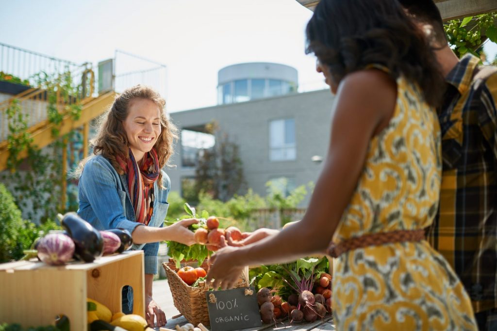 A smiling vendor hands fresh produce to a customer at an outdoor farmers market stand labeled “100% Organic.”