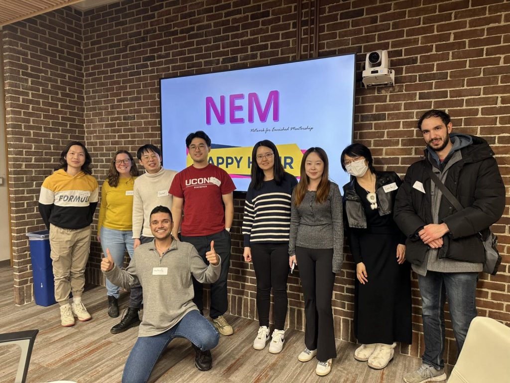 A group of people is posing and smiling together indoors in front of a screen that reads “NEM Happy Hour,” suggesting a friendly social or networking event.