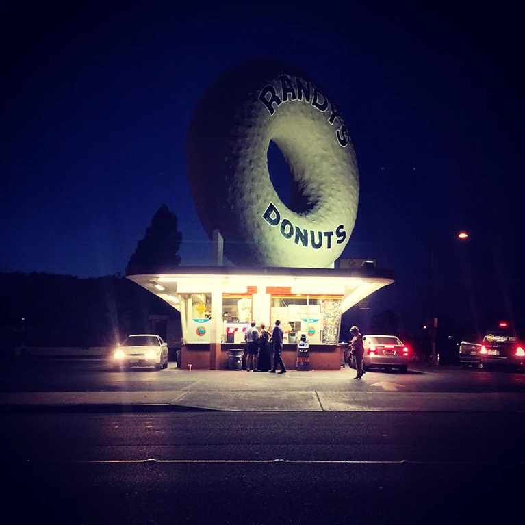 A brightly lit donut shop at night features a giant donut sign on its roof, with people and cars gathered around the storefront.
